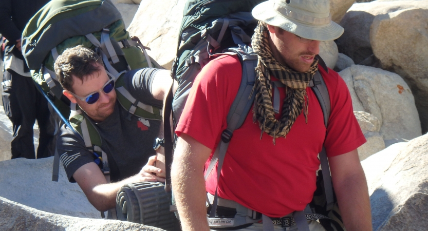 One person helps adjust the backpack of another amidst large rocks.